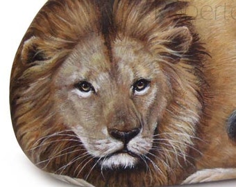 May include: A painted rock depicting a lion with a brown mane and a light brown body. The lion is lying down with its head resting on its paws. The rock is painted realistically and the lion's eyes are open and looking directly at the viewer.