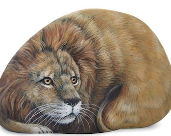 May include: A hand-painted stone depicting a lion with a realistic, detailed style. The lion has a brown and tan coat with a white chest and a long, flowing mane. The lion's eyes are open and looking directly at the viewer.