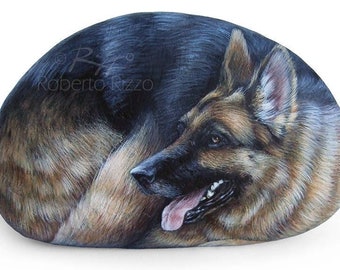 May include: A hand-painted rock depicting a German Shepherd dog lying down with its head turned to the side. The dog's fur is brown and black, and its eyes are brown. The rock is painted realistically, with details such as the dog's fur, eyes, and tongue.