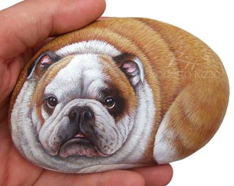 May include: Hand-painted rock art depicting a brown and white bulldog with a serious expression. The rock is oval-shaped and the bulldog is painted realistically with details like wrinkles and fur.