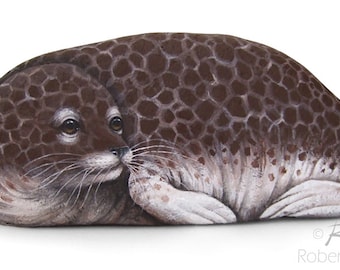May include: A hand-painted rock art piece depicting a brown and white seal with a spotted pattern. The seal is curled up in a sleeping position with its eyes closed.