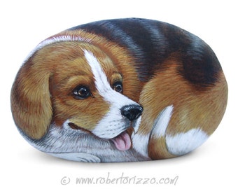 May include: Hand-painted rock art depicting a beagle dog with brown, black, and white fur. The dog is lying down with its head resting on its paws and its tongue sticking out. The rock is smooth and oval-shaped.