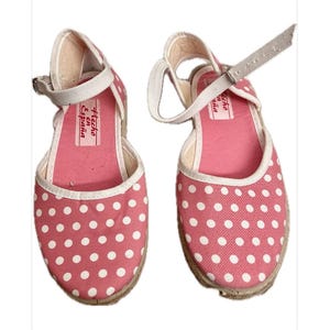 Puede incluir: Par de sandalias espadrillas rosas con lunares blancos y ribete blanco. Las sandalias tienen una plantilla rosa y una correa de tobillo blanca con hebilla plateada. Las suelas son de yute tejido. El texto "Hecho en España" es visible.