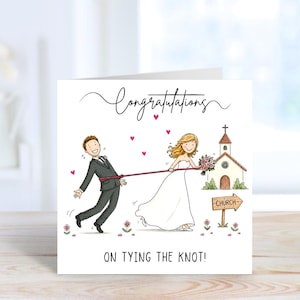 Könnte beinhalten: Quadratische Hochzeitskarte mit Cartoon-Illustration. Eine Braut in einem weißen Kleid zieht einen Bräutigam im Anzug mit einem Seil in Richtung einer Kirche. Die Karte trägt die Aufschrift "Congratulations" und "On tying the knot!" mit rosa Herzen.