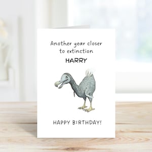 Peut inclure: Une carte de v&oelig;ux blanche avec une illustration de dodo gris au centre. Le texte indique Another year closer to extinction HARRY HAPPY BIRTHDAY en lettres noires. La carte est plac&eacute;e verticalement sur une surface en bois.