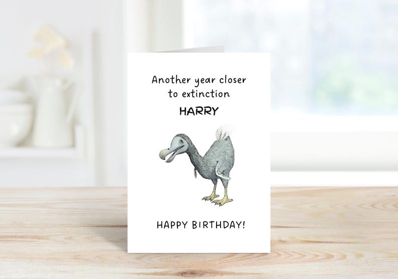 Peut inclure: Une carte de v&oelig;ux blanche avec une illustration de dodo gris au centre. Le texte indique Another year closer to extinction HARRY HAPPY BIRTHDAY en lettres noires. La carte est plac&eacute;e verticalement sur une surface en bois.