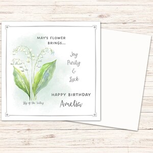 Puede incluir: Tarjeta de cumplea&ntilde;os con una ilustraci&oacute;n de acuarela de lirios de los valles. La tarjeta incluye el texto: "May's Flower Brings... Joy, Purity & Luck. Happy Birthday Amelia." Se incluye un sobre blanco.