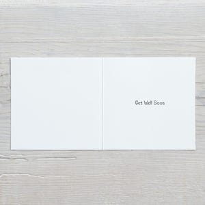 May include: An open white greeting card with the message "Get Well Soon" printed in black. The card is displayed on a light grey wooden surface.