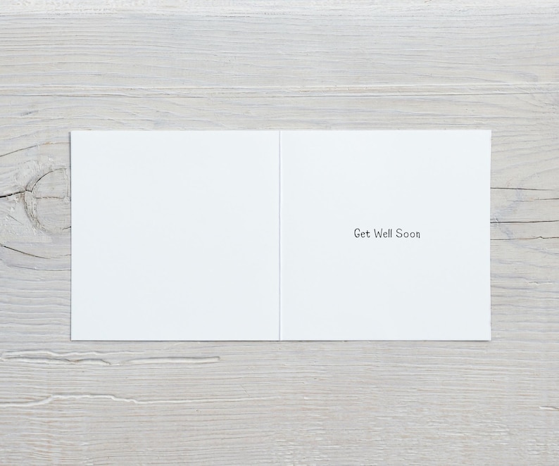 May include: An open white greeting card with the message "Get Well Soon" printed in black. The card is displayed on a light grey wooden surface.