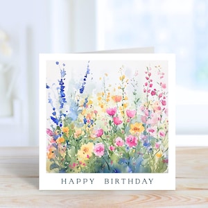 Peut inclure: Une carte de vœux carrée présentant une illustration à l'aquarelle d'une prairie de fleurs sauvages aux tons bleus, jaunes et roses. Le texte "HAPPY BIRTHDAY" est imprimé en police noire sans empattement au bas de la carte.