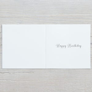 K&ouml;nnte beinhalten: Ge&ouml;ffnete wei&szlig;e Geburtstagskarte mit dem Schriftzug "Happy Birthday" in eleganter Schrift. Die Karte liegt auf einem Hintergrund aus hellgrauen Holzplanken.