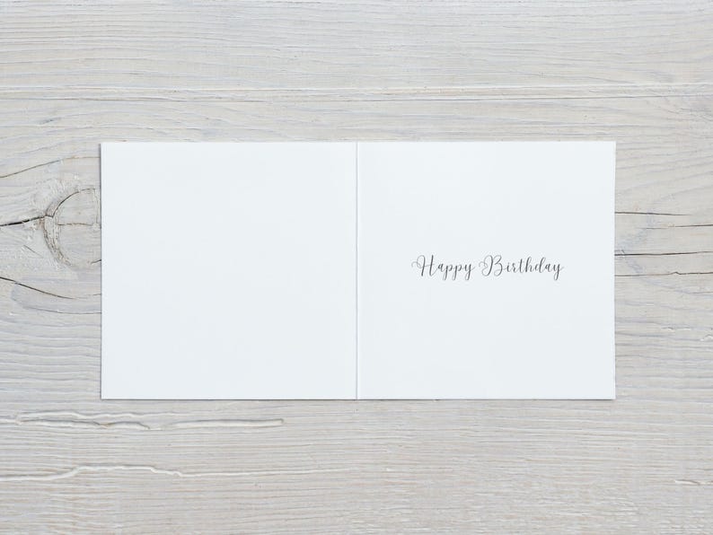 K&ouml;nnte beinhalten: Ge&ouml;ffnete wei&szlig;e Geburtstagskarte mit dem Schriftzug "Happy Birthday" in eleganter Schrift. Die Karte liegt auf einem Hintergrund aus hellgrauen Holzplanken.