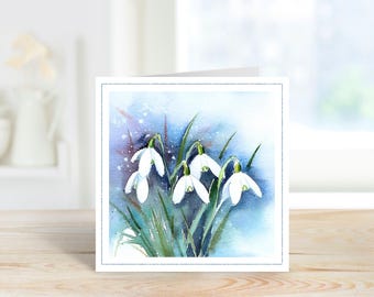 May include: A square greeting card featuring a watercolor painting of white snowdrop flowers with green stems and leaves. The background is a soft, blended wash of blue and purple tones with white paint splatters, framed by a thin border.
