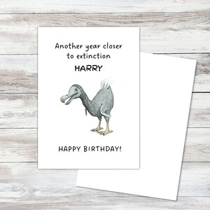 Peut inclure: Une carte de v&oelig;ux blanche illustr&eacute;e d'un dodo gris. Le texte indique : Another year closer to extinction, HARRY, HAPPY BIRTHDAY ! La carte est rectangulaire avec une inscription noire et un dessin d&eacute;taill&eacute; de l'oiseau disparu.
