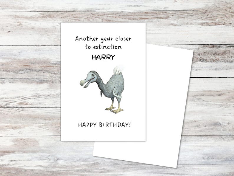 Peut inclure: Une carte de v&oelig;ux blanche illustr&eacute;e d'un dodo gris. Le texte indique : Another year closer to extinction, HARRY, HAPPY BIRTHDAY ! La carte est rectangulaire avec une inscription noire et un dessin d&eacute;taill&eacute; de l'oiseau disparu.