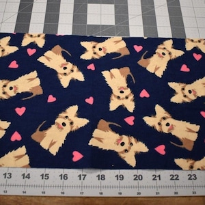 May include: Navy blue fabric featuring a repeating pattern of tan and brown cartoon dogs and pink hearts. The fabric is measured with a ruler marked in inches. The dogs have smiling faces and are in various poses.