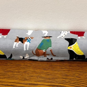May include: A gray fabric door draft stopper featuring a repeating pattern of various dog breeds. Some dogs wear yellow vests, green sweaters, or red coats. The dogs are set against a light gray background.