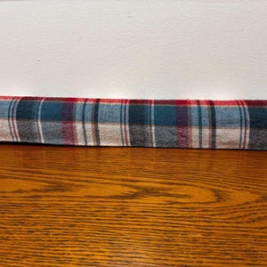 May include: A long, cylindrical draft stopper featuring a classic plaid pattern in shades of red, dark blue, white, and grey. The fabric has a soft, textured appearance, and the object is positioned horizontally across the frame in a straight line.