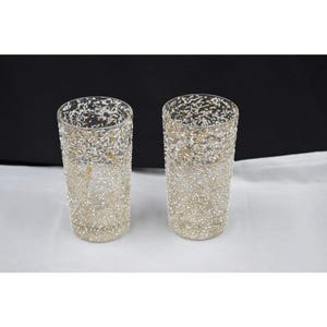 May include: Two clear glass tumblers featuring a textured, speckled exterior pattern in white and metallic gold. The cylindrical drinking glasses have a straight-sided design with a consistent, dense distribution of flecks covering the entire surface.