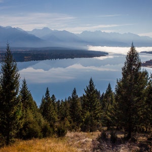May include: A scenic view of a lake with mountains in the background. The lake is calm and reflects the sky and clouds. There are tall evergreen trees in the foreground.