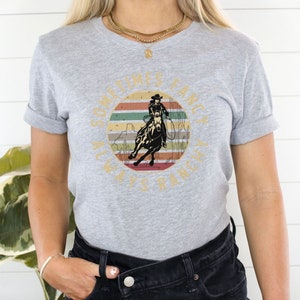 Op de afbeelding: Lichtgrijs t-shirt met een cirkelvormig ontwerp van een cowboy op een paard tegen een zonsondergang. De tekst "SOMETIMES FANCY, ALWAYS RANCHY" staat rond de afbeelding. De mouwen zijn opgerold. Casual kleding.