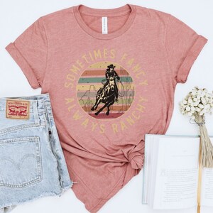 Op de afbeelding: Een stoffig roze t-shirt met opgerolde mouwen en de tekst "Sometimes Fancy, Always Ranchy" rond een afbeelding van een cowboy op een paard die een stier vangt. Lichtblauwe spijkerbroekshorts liggen naast het shirt.
