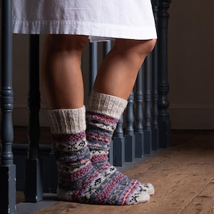 May include: A person wearing patterned wool socks with a cream-coloured cuff. The socks feature a geometric design in shades of red, blue, purple, and black. The person is standing on a wooden floor near a blue banister.