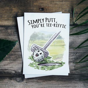 May include: A white card with a watercolor illustration of a golf ball going into a hole. The text on the card reads "Simply putt, you're tee-riffic."