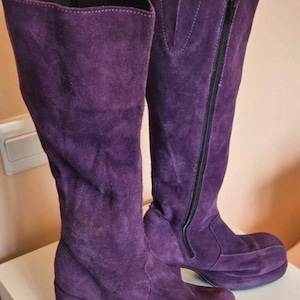 Puede incluir: Un par de botas altas de ante morado. Las botas tienen tacón ancho, suela de plataforma y cierre de cremallera lateral. Están hechas de un material suave y texturizado. Altura del tacón: aprox. 7 cm.
