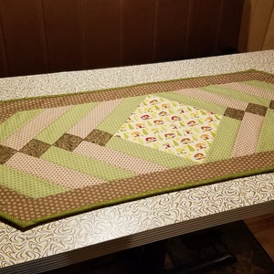 May include: A green, brown and white quilted table runner with a diamond pattern. The centre diamond features a print of trees and mushrooms.