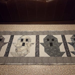 May include: A rectangular quilted table runner featuring four ghost figures in shades of gray, black, and white. The ghosts are separated by dark patterned fabric strips, and the entire piece is bordered by a gray and white striped fabric frame.