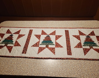 May include: A quilted Christmas table runner with a repeating pattern of red and white starbursts and Christmas trees. The trees are made of green, brown, and teal fabric. The runner is approximately 30 cm wide and 102 cm long.