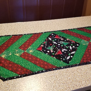 May include: A green and red quilted table runner with a black diamond-shaped centre featuring a martini glass pattern. The runner has a black border.