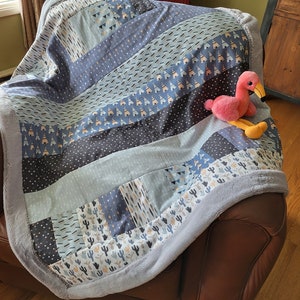 May include: A blue and gray patterned baby blanket with a plush border. The blanket is folded on a brown leather couch with a pink flamingo toy on top.