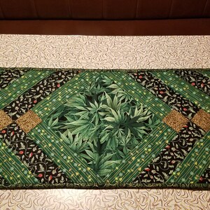 May include: A green and gold table runner with a diamond pattern. The centre of the runner features a green leafy design.