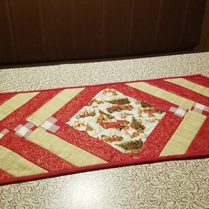 May include: A red and gold table runner with a diamond-shaped centre featuring a Christmas-themed print of red trucks carrying Christmas trees. The runner has a white and red checked pattern on the sides.
