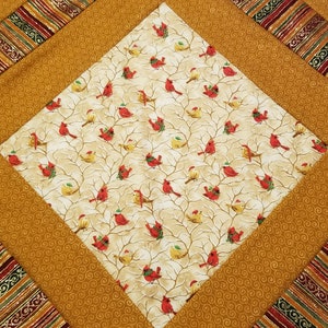May include: A quilted throw featuring a patchwork design with a central square of red robins on a cream background. The surrounding squares are a solid gold colour with a swirling pattern and a red and gold striped pattern.