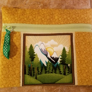 May include: A yellow fabric zippered pouch with a green zipper pull and a green and white polka dot ribbon. The pouch features an embroidered image of a mountain scene with a sun, trees, and snow-capped peaks.
