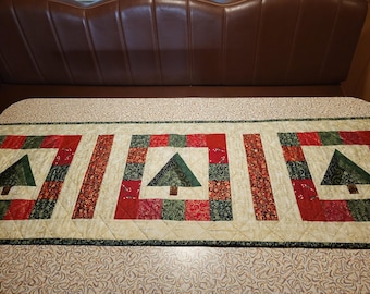 May include: A quilted Christmas table runner with a repeating pattern of green Christmas trees and red and green squares. The runner has a cream-coloured background and is bordered with a green trim. The runner is approximately 30 cm wide.