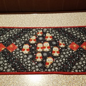 May include: A black and red quilted table runner with a Christmas theme. The runner features a repeating pattern of snowflakes and holly leaves. There are also several small, white owls with red hats and scarves.