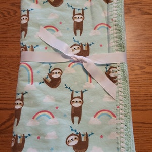 May include: A green baby blanket with a white crocheted border. The blanket features a repeating pattern of sloths hanging from branches with rainbows and stars on a light blue background.