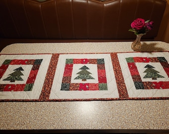 May include: A quilted Christmas table runner with three square panels, each displaying a green Christmas tree appliqué. The panels are bordered by red and green patchwork and a patterned edge. The runner is on a patterned table.