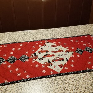 May include: A red and black quilted table runner with a snowflake pattern and a design of red trucks and Christmas trees. The runner has a black and white trim.