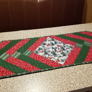 May include: A green and red quilted table runner with a diamond-shaped centre featuring a winter scene with trees and red trucks. The runner has a scalloped edge.