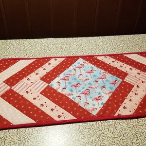 May include: A red and white quilted table runner with a diamond-shaped centre featuring a pink flamingo pattern on a blue background. The runner has a pink border and is made of various fabrics.