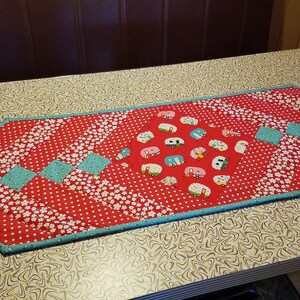May include: A quilted table runner with a red backdrop, white floral and polka dot patterns. The centre displays camper illustrations. Turquoise squares and trim complete the design. It measures approximately 102 cm in length.