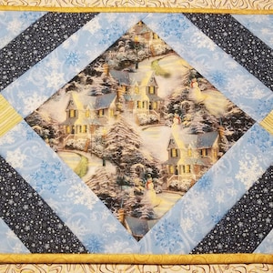 May include: A quilted table runner with a diamond-shaped centre featuring a winter scene of houses and snow-covered trees. The runner is made of blue, yellow, and white fabric with a snowflake pattern.