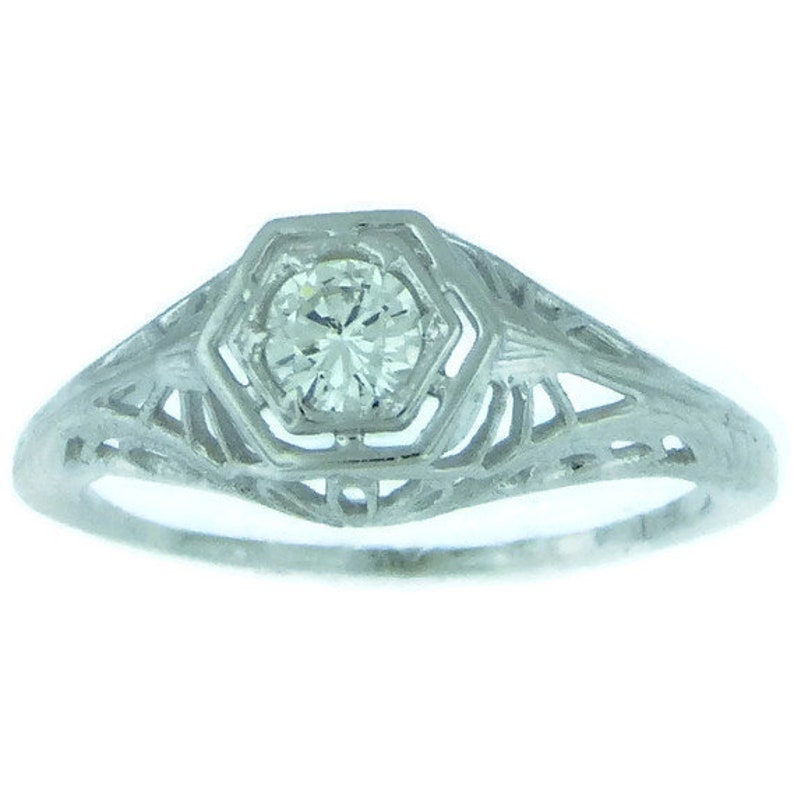 May include: A delicate, filigree engagement ring with a single round diamond set in a hexagonal bezel. The ring is made of white gold and has a vintage style.