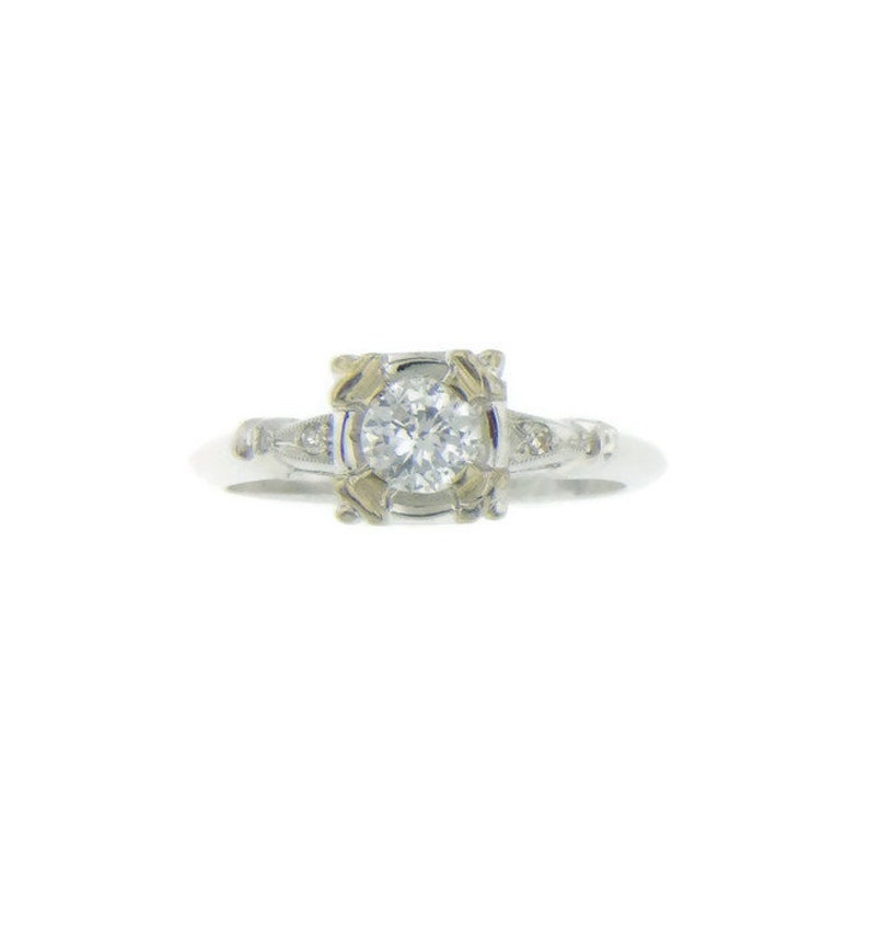 May include: A silver ring with a round, clear diamond set in a square frame. The frame has a gold-toned design. The ring has smaller diamonds on the band.