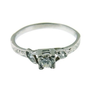 May include: A silver ring with a single diamond and two smaller diamonds on the band. The band is decorated with an intricate design.